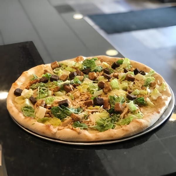 Caesar salad pizza with chicken, croutons, and lettuce on a dark table.