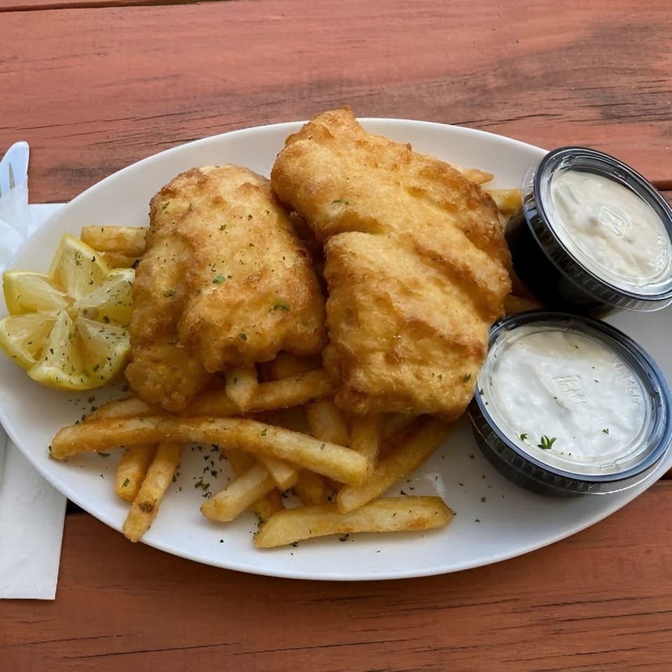 Golden-brown fish and chips with lemon wedges and two containers of tartar sauce on a white plate.