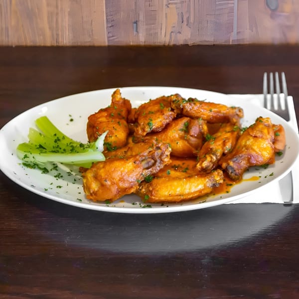 A white plate of buffalo chicken wings with celery sticks and parsley on a dark wooden table.