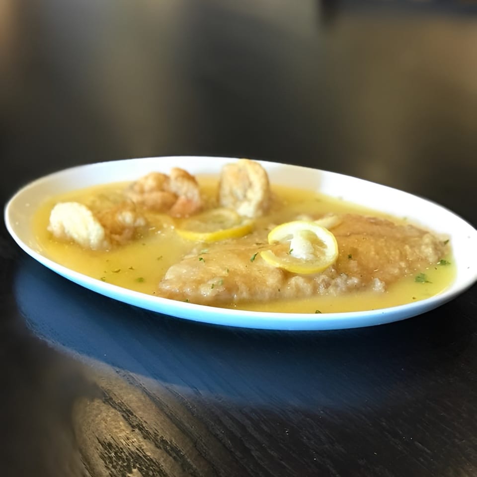 Chicken piccata with lemon slices and parsley in a white oval dish on a dark table.