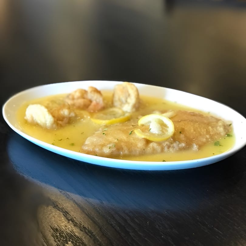 Chicken piccata with lemon slices and parsley in a white oval dish on a dark table.